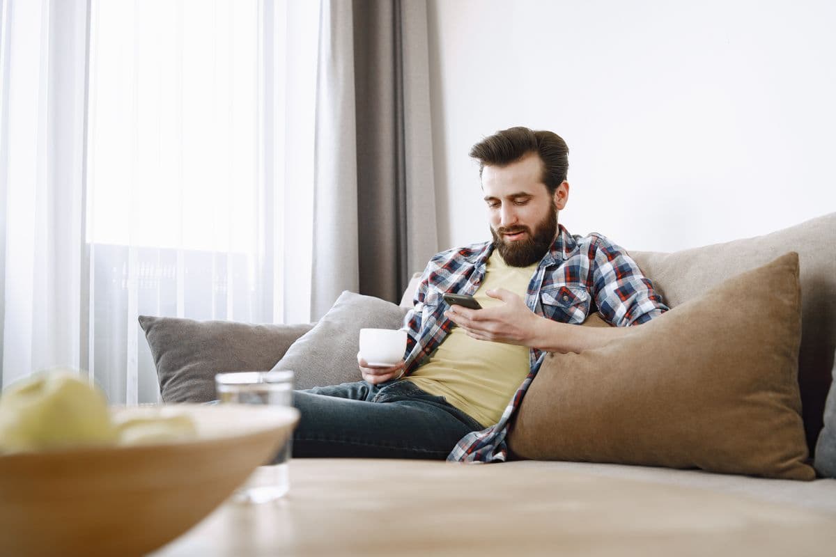 A man relaxing on a sofa with a coffee, browsing room listings on his phone