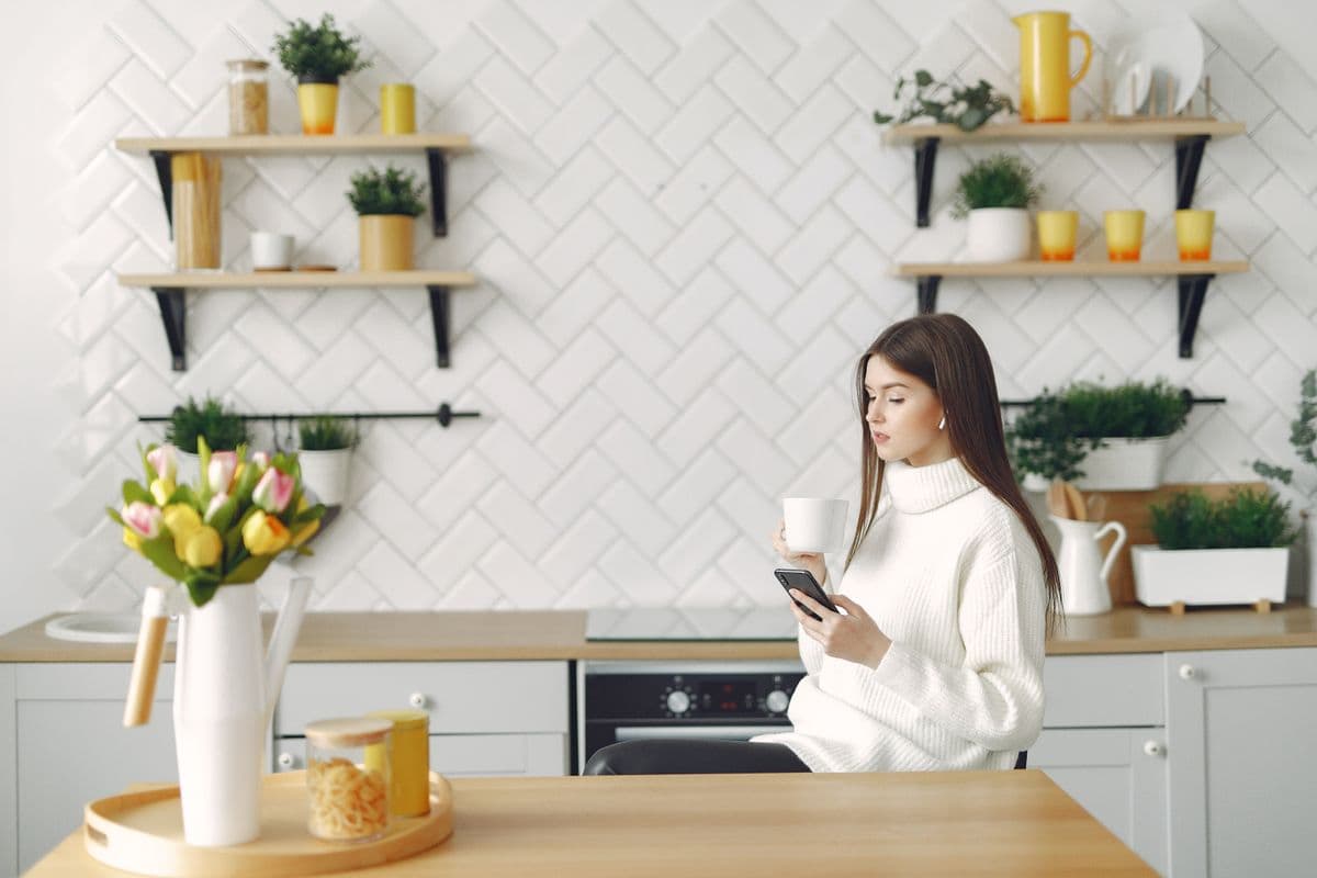 A woman with a coffee browsing room listings on her phone in a bright kitchen